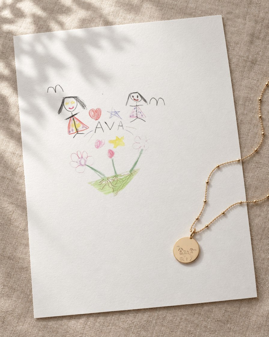 A child's crayon drawing of two girls, a heart and flowers on paper, with the same drawing engraved on a gold pendant resting on the page